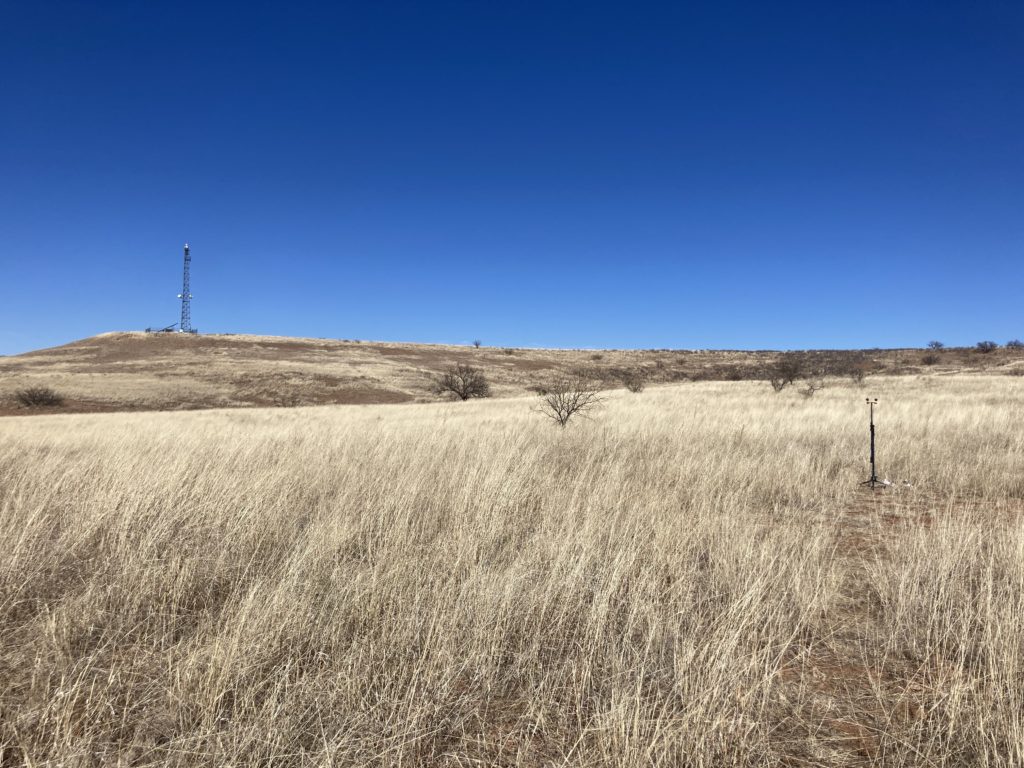 long shot of grassy field, mic stand small in mid-ground to the right, DHS Integrated Fixed tower on a hill way in the foreground in the left