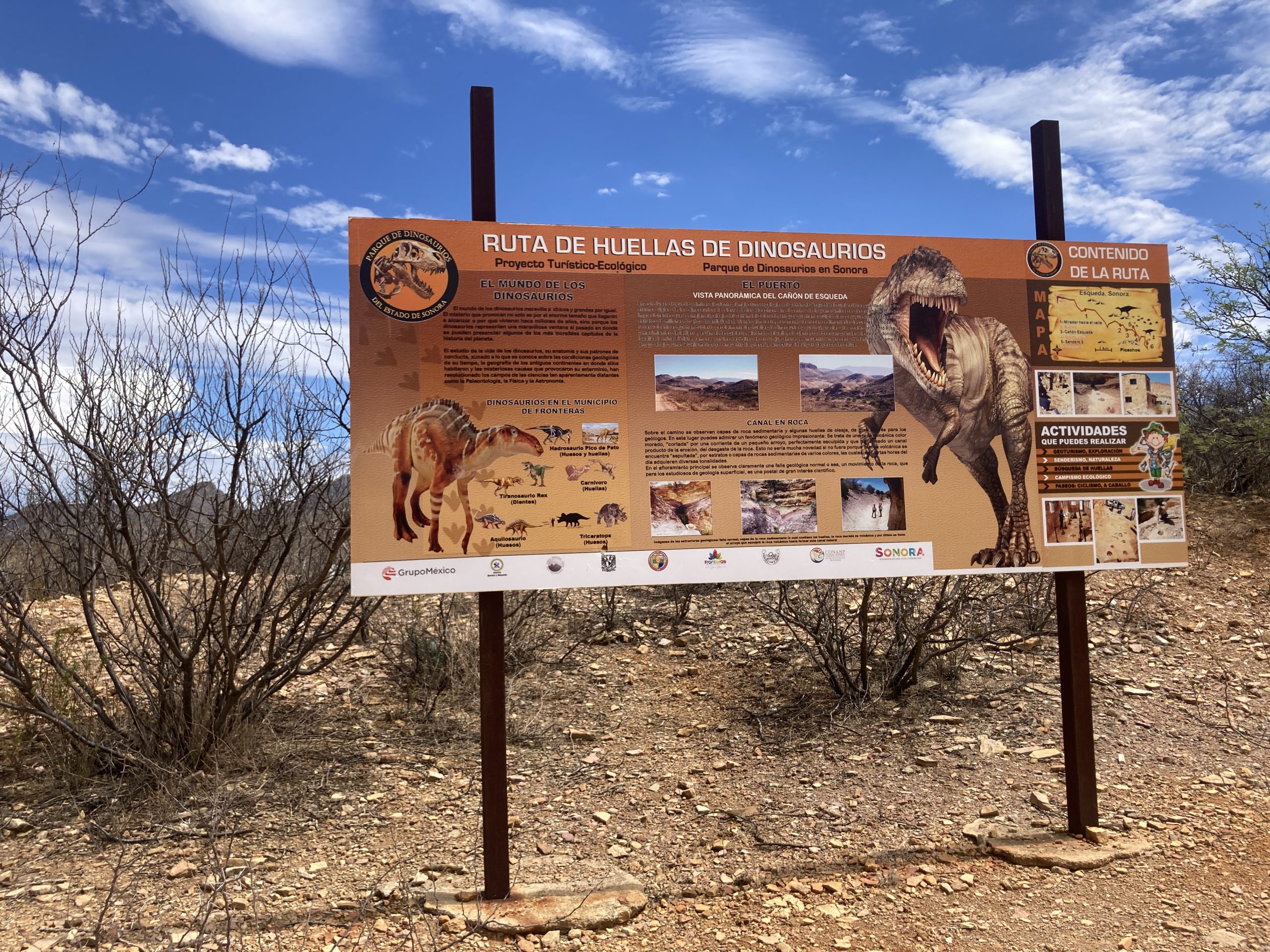 A sign, in spanish, for "La Ruta de los Dinosaurios". It shows pictures of various dinosaurs and descries how the area was 70 million years ago. In the background is a blue sky with clouds and a desert landsacpe