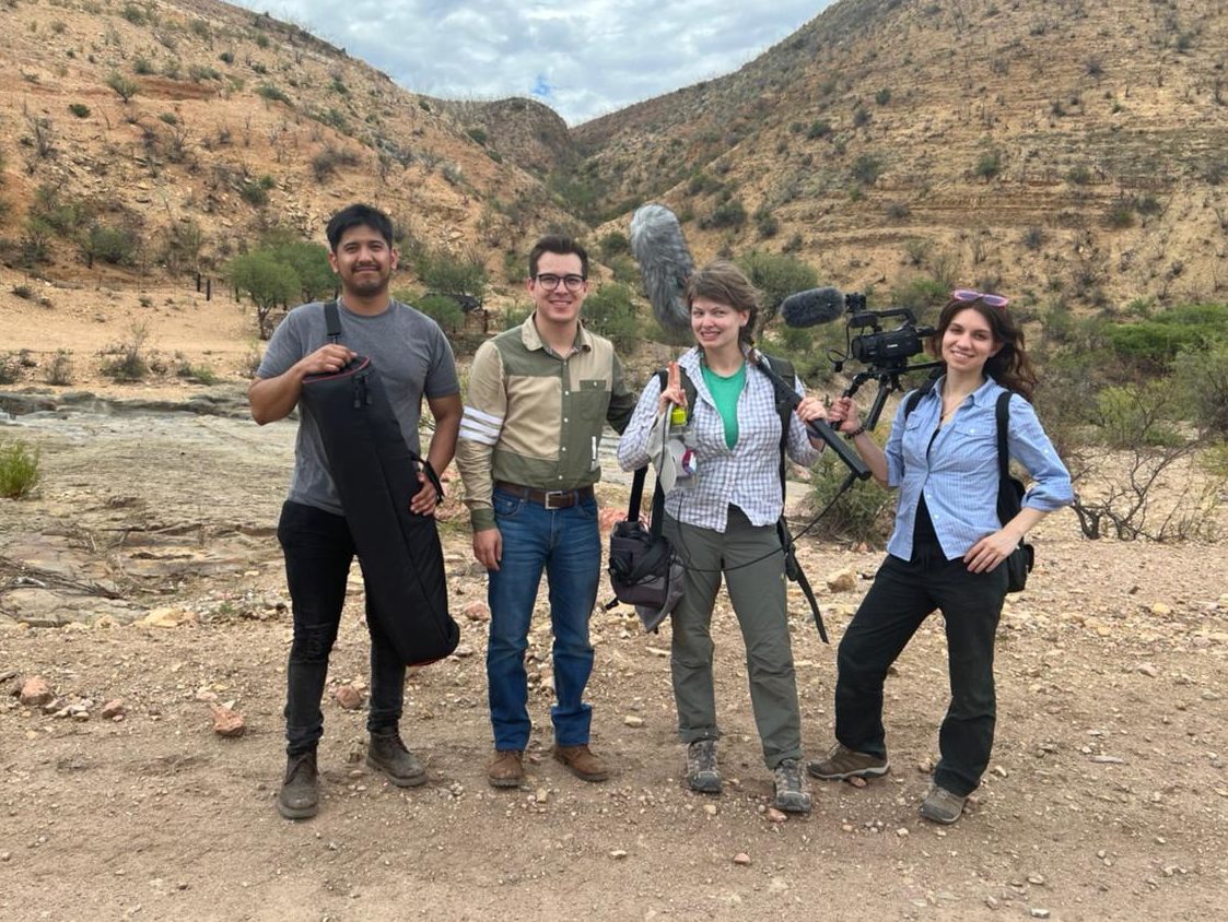 A photo of four people standing in a hilly, desert landscape. They are smiling at the camera and holding video and audio equipment