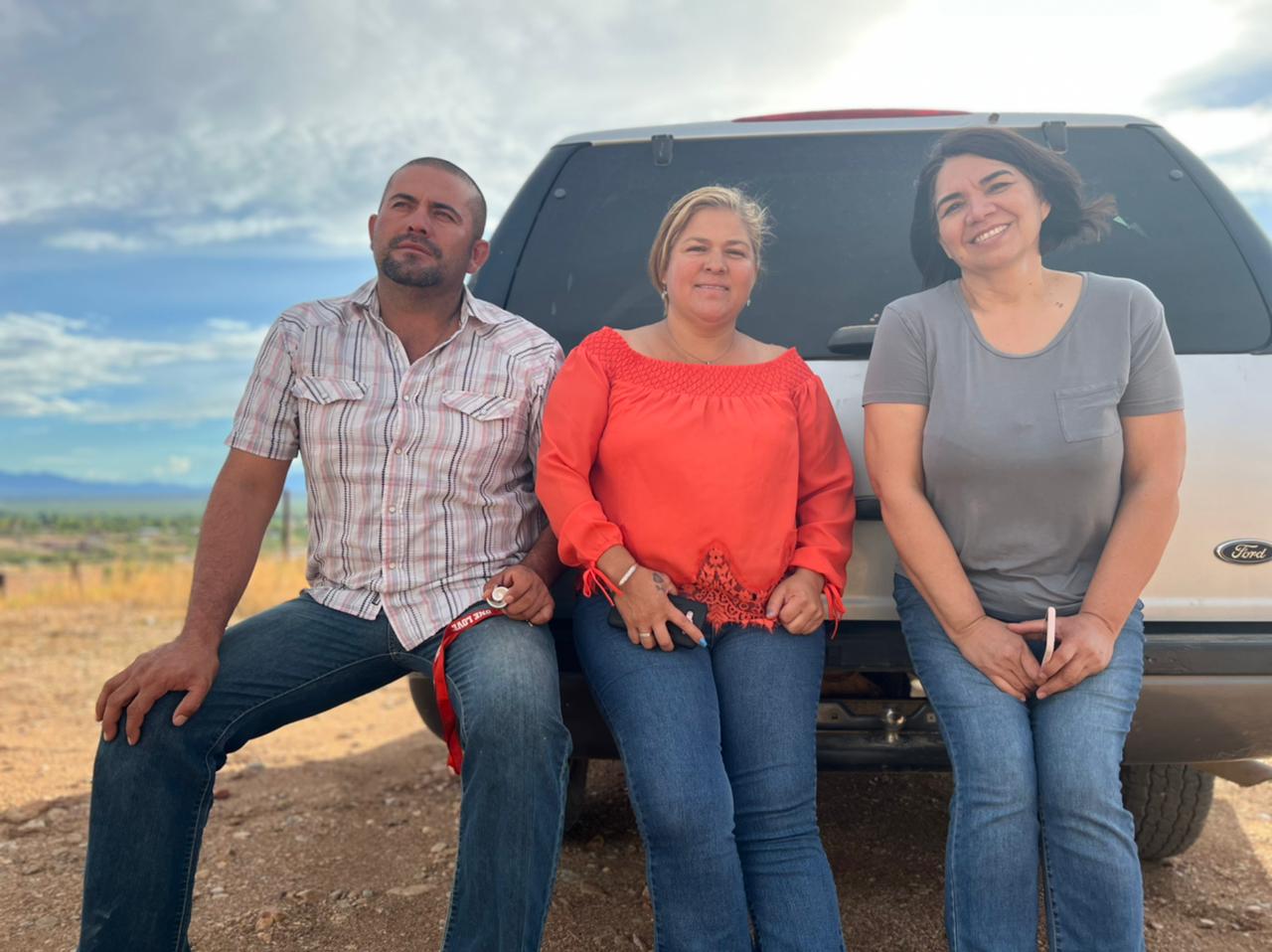 Three people are sitting on the bumper of an SUV. They are looking beautiful for the camera